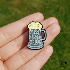 a hand holding a pin with a beer on it that says raise a glass to freedom