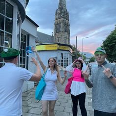 three people standing in front of a building with a clock tower behind them and one woman holding up her hand