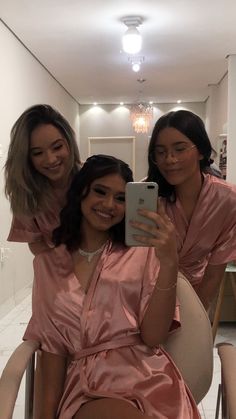 three women in pink robes are taking a selfie with a cell phone while sitting on a chair