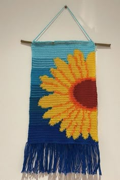 a crocheted sunflower hanging on a wall with blue and yellow fringes