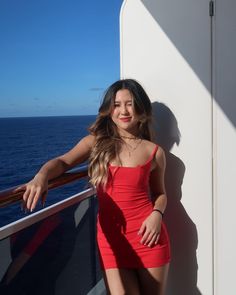 a woman in a red dress leaning against a wall next to the ocean on a cruise ship