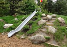 a slide that is sitting in the grass next to some rocks and trees on top of a hill