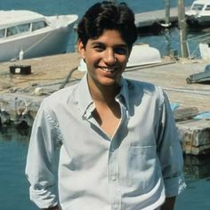 a young man standing in front of a body of water with boats docked behind him