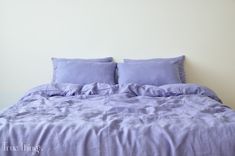 a bed with purple sheets and pillows on top of it in a white walled room