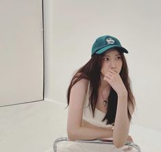 a young woman sitting on top of a glass table next to a white wall and wearing a blue hat