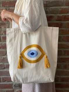 This mustard yellow evil eye boho tote bag made of Turkish Buldan cotton fabric with hidden pocket inside. Hand crocheted evil eye pattern is made of %100 cotton yarn. Hand crocheted evil eye sewn on a natural Turkish Buldan cotton fabric bag  with a sewing machine. Double layer cotton tote will be a great helper in your everyday use. Dimensions & Info: Width: 17 inches / Height 18.5 inches / Bottom depth 3.5 inches / Strap length  26.5 inches Tote bag has two pockets inside:  5x 7.5 inches / 3x White Handmade Bohemian Canvas Bag, Handmade Bohemian Canvas Shoulder Bag, Bohemian Cotton Canvas Shoulder Bag, Handmade Beige Cotton Beach Bag, Bohemian Beige Cotton Shoulder Bag, Beige Bohemian Cotton Bag, Beige Cotton Bohemian Bag, Bohemian Beige Cotton Bag, Bohemian Cotton Crochet Bag For Everyday Use