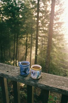 two cups of coffee sitting on top of a wooden deck in the woods with trees