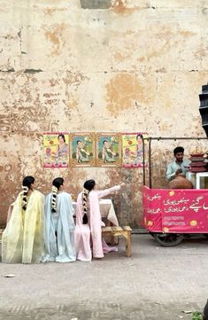 four women in sari are sitting on a bench near a street vendor's cart