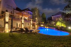 an outdoor swimming pool surrounded by lawn furniture and landscaping lights at night in front of a house