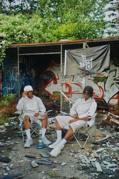 two men sitting on folding chairs in front of graffiti covered walls and trees, one with his head down