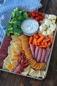 a platter filled with meat, cheese, crackers and veggies next to a bowl of dip