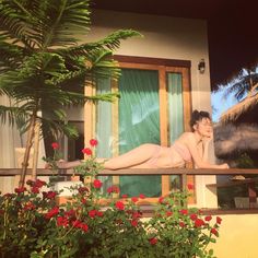 a naked woman laying on top of a balcony next to flowers and palm trees in front of a house