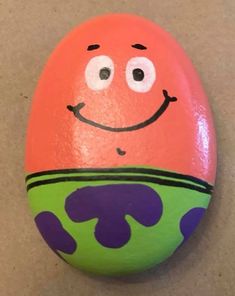 an orange and green painted rock with a smiling face on it's side, sitting on a tan surface