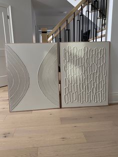 two framed art pieces sitting on top of a hard wood floor next to a stair case