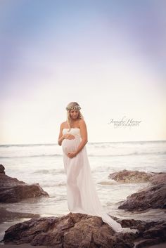 a pregnant woman in a white dress standing on rocks by the ocean with her belly wrapped around her waist