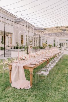 a long table set up with clear chairs and pink linens on the grass in front of a large building