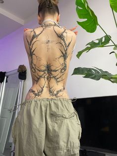 a woman with tattoos on her back standing in front of a plant and looking at the television