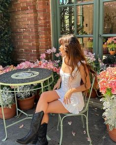 a woman sitting at a table in front of some flowers and potted plants with her legs crossed