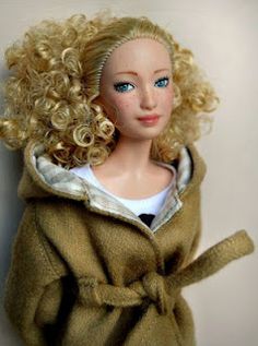 a doll with blonde hair and blue eyes wearing a brown coat on a white background