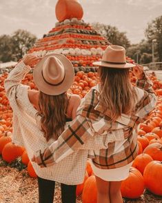 two women standing next to each other in front of pumpkins at a field with one woman wearing a hat and the other holding her arms behind her back