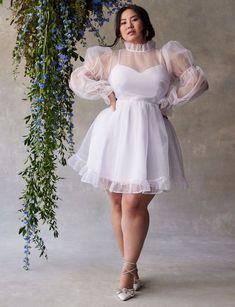 a woman in a short white dress standing next to a flower bush with her hands on her hips