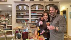 a man and woman standing next to each other holding glasses in front of shelves filled with bottles