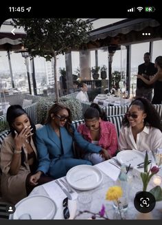 three women sitting at a table with plates and drinks in front of them, talking on the phone