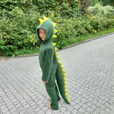 a young boy in a green dinosaur costume standing on a brick walkway with trees and bushes behind him