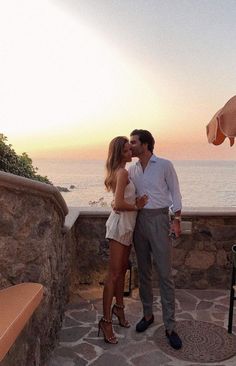 a man and woman standing next to each other in front of the ocean at sunset