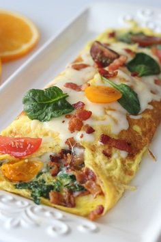 an omelet with bacon, spinach and tomatoes on a white plate next to sliced oranges