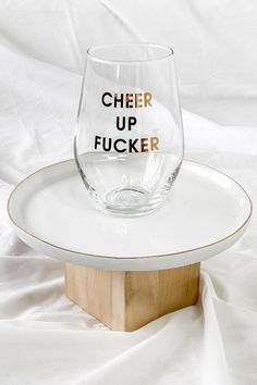 a clear wine glass sitting on top of a white plate with the words cheer up focker