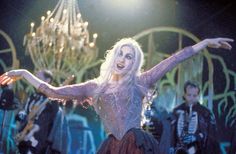 a woman in costume dancing on stage with other people behind her and chandelier hanging from the ceiling