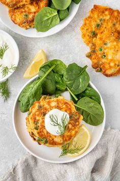 two white plates topped with crab cakes and spinach leaves next to lemon wedges