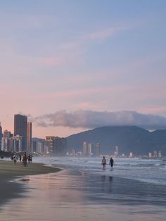 people are walking on the beach in front of some tall buildings and skyscrapers at sunset