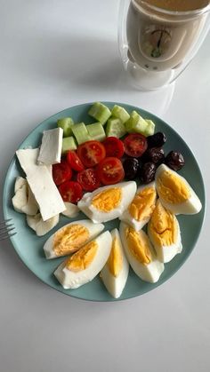 a plate with hard boiled eggs, tomatoes, cucumbers and olives next to a cup of coffee