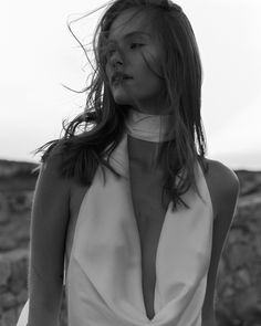 a woman with long hair wearing a halter top and white pants standing in front of rocks