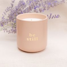 a pink candle with the words be still on it next to some lavender flowers and a white cloth