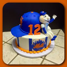 a baseball themed cake with an orange and blue hat on it's head, sitting on a tile surface