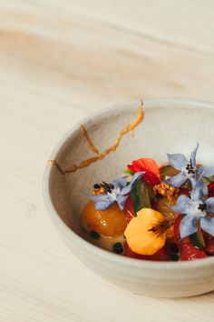 a white bowl filled with fruit and flowers on top of a wooden table next to a fork