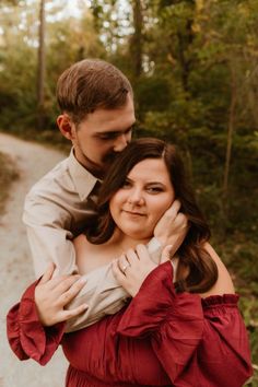 a man and woman hugging each other in the woods on a dirt road surrounded by trees