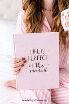 a woman in pajamas holding a pink notebook with the words life is perfect in this moment