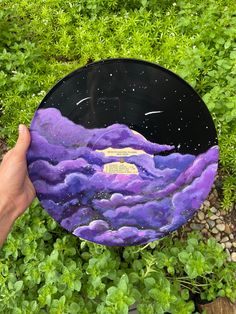 a hand holding up a purple and black plate with clouds painted on it in the grass