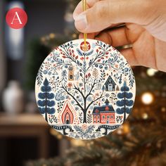 a hand holding a christmas ornament in front of a tree with houses and trees on it