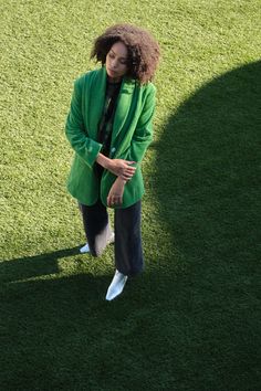 a woman standing in the grass wearing a green jacket and white sneakers with her hands on her hips