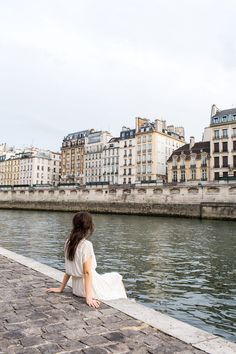 a woman sitting on the side of a river next to tall buildings and looking at the water