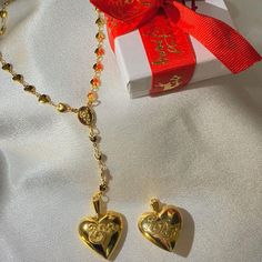 two gold heart shaped pendants on a chain next to a red ribbon and gift box