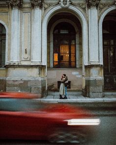 two people standing in front of an old building with cars passing by on the street