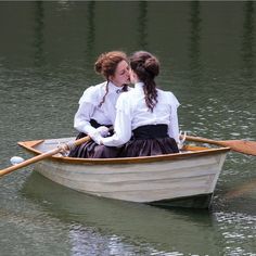 two women are sitting in a rowboat on the water, one wearing a white shirt and black skirt