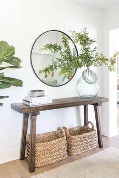 a wooden table topped with a vase filled with green plants next to a mirror and wicker baskets