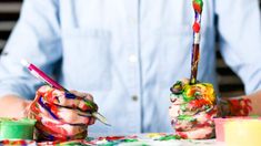 two hands holding paintbrushes and paints on a table